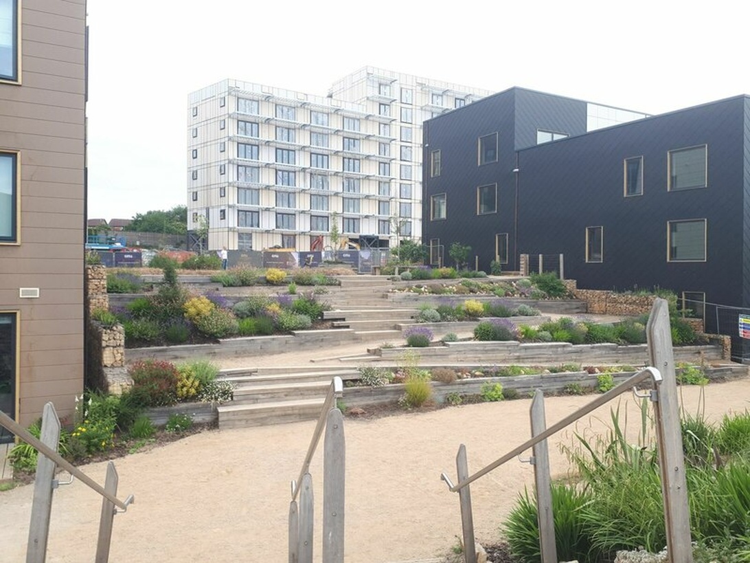 Ett modernt bostads- eller blandat utvecklingsområde med terrasserad landskapsarkitektur och planterade ytor mellan byggnaderna. Integrationen av grönska och terrasserade trädgårdar antyder principer för hållbar design, vilket ofta går hand i hand med energieffektivitet.